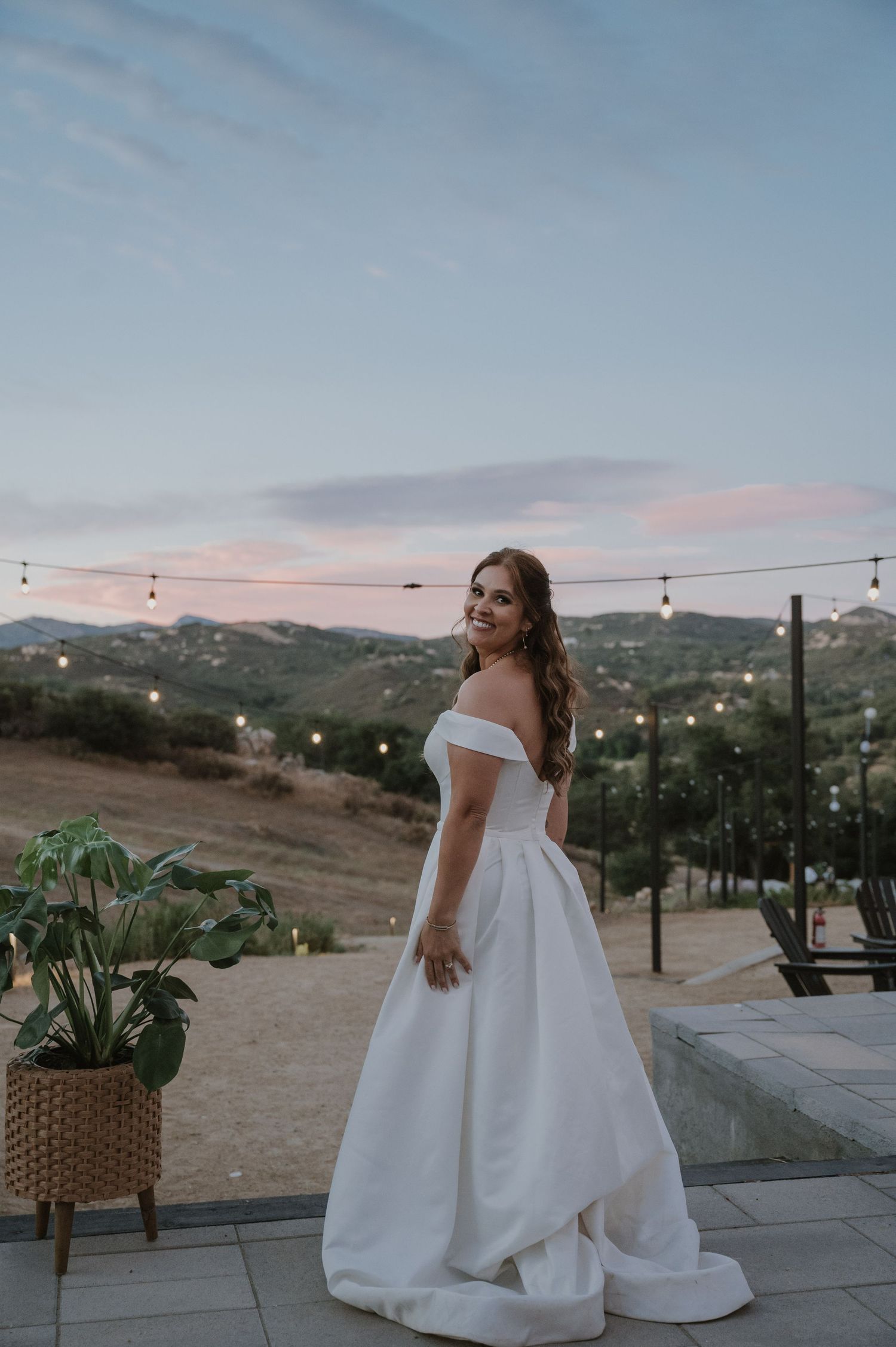 Bride in off-shoulder white ball gown smiling at sunset with string lights and rolling hills backdrop.