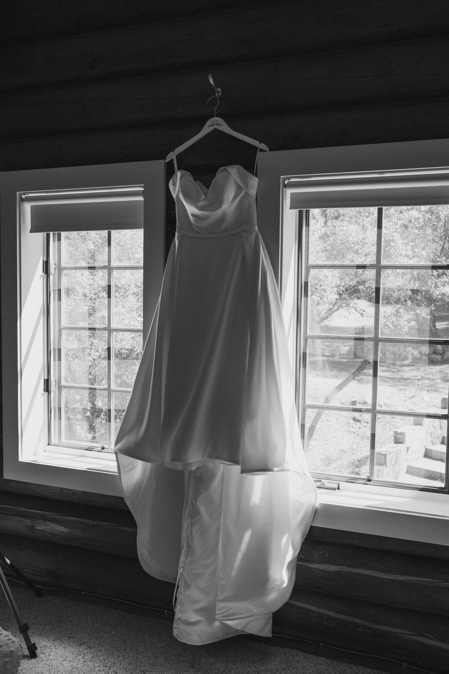 Elegant strapless wedding gown hanging on a hanger in front of bright windows in black and white.