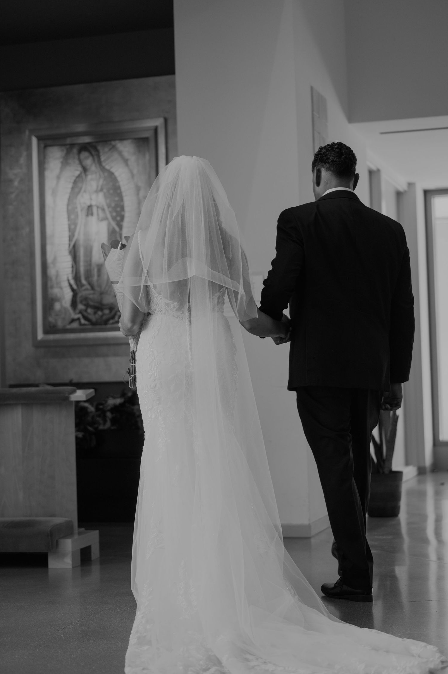 Black and white photo of bride and groom walking hand in hand in a church with Our Lady of Guadalupe artwork.