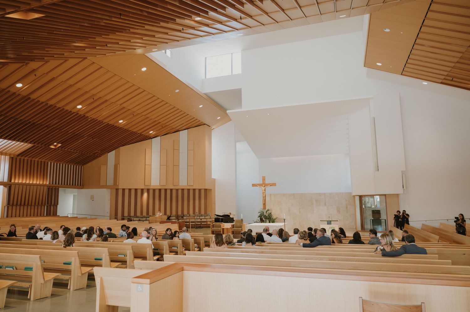 Modern church interior with wooden ceiling, pews filled with wedding guests, and a cross at the altar during a ceremony.