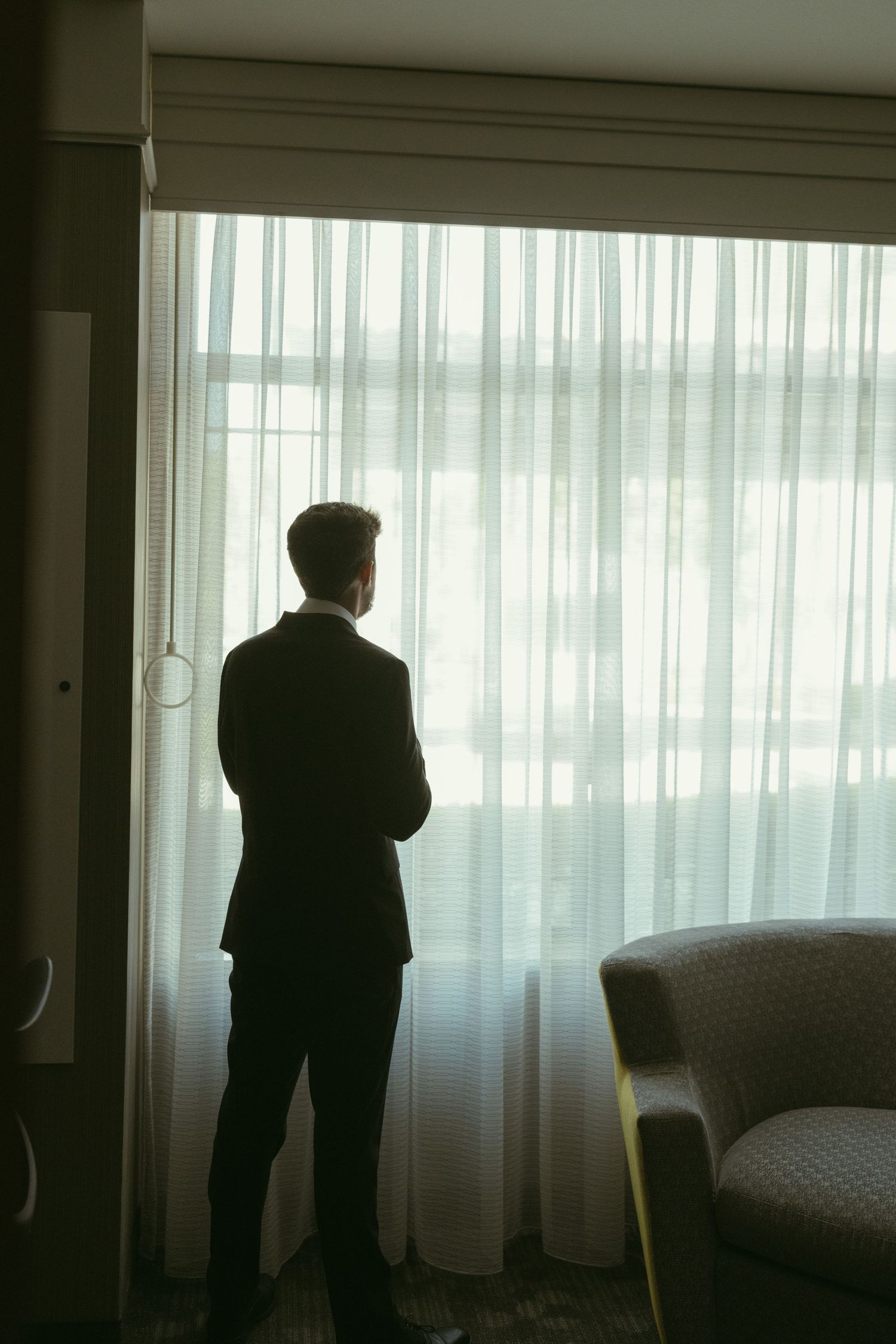 A groom in a dark suit stands contemplatively before sheer curtains in a hotel room before his wedding.