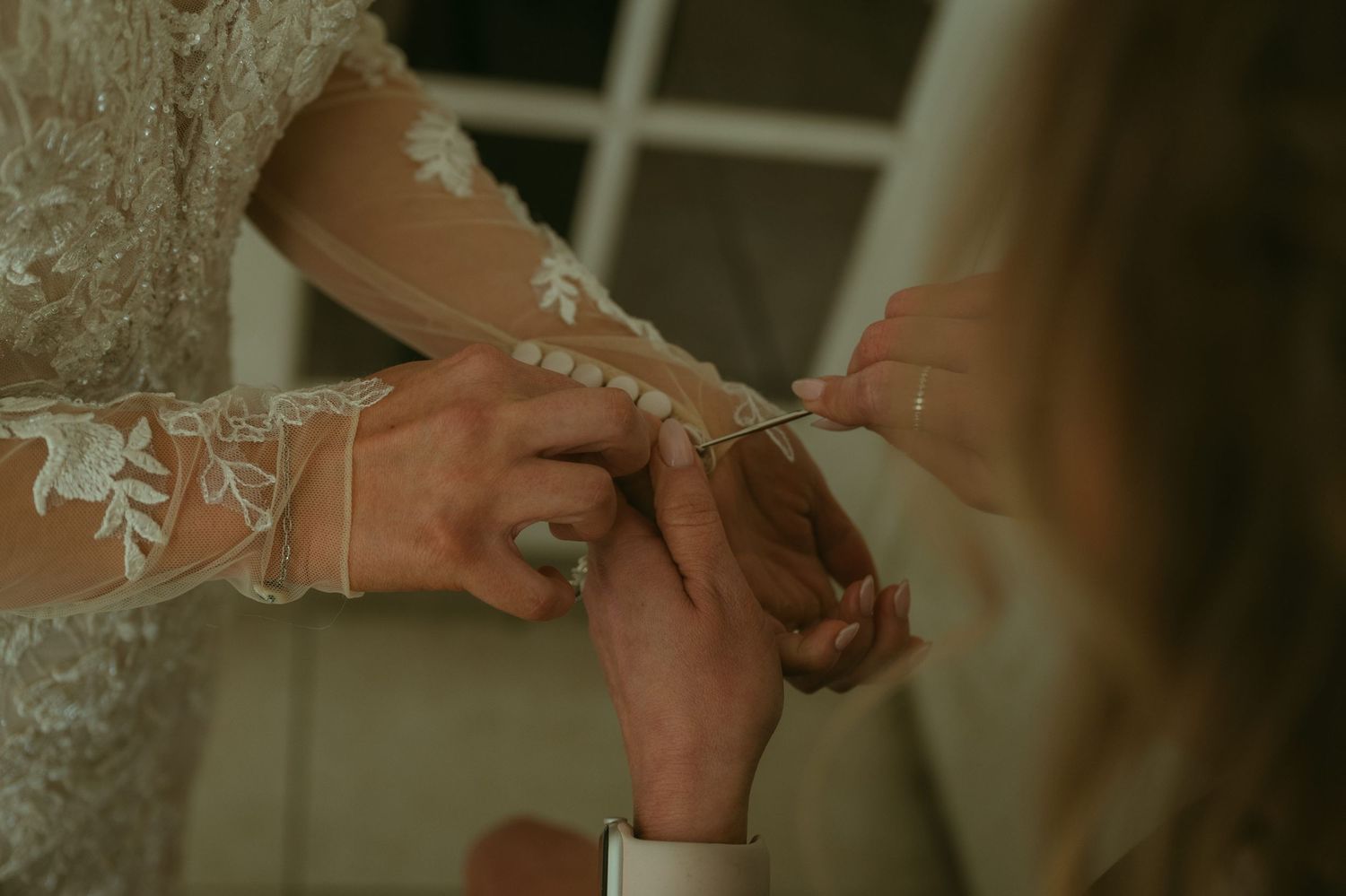 Close-up of a bride in lace wedding dress having her sleeve buttoned by a helper during wedding preparations.