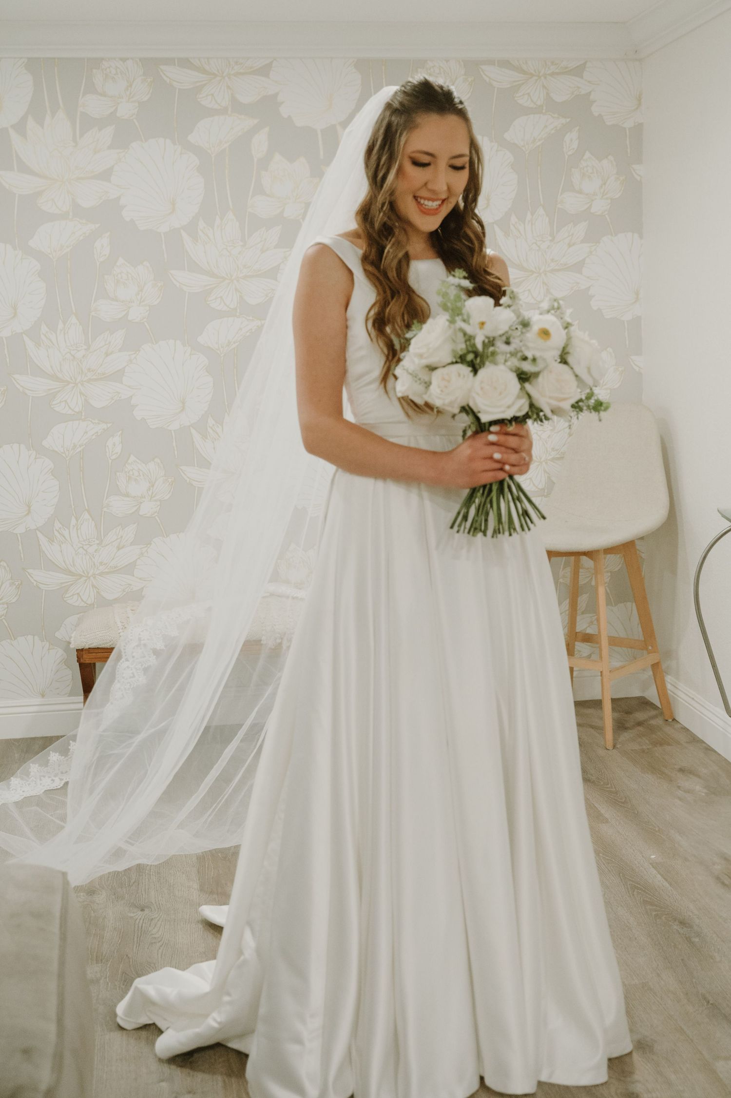 Radiant bride in sleeveless satin gown with cathedral veil holding white rose bouquet indoors.