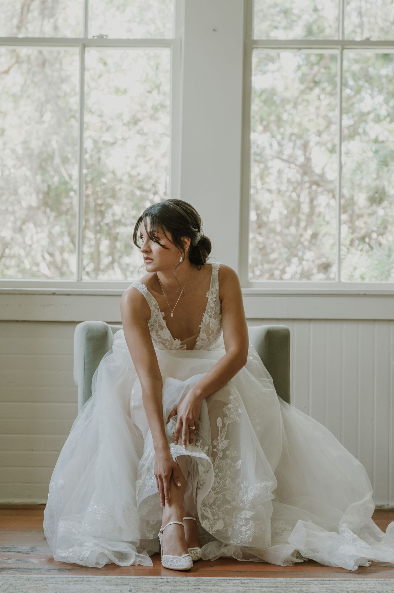 Bride in a lace wedding gown sits in a chair by a window, adjusting her white heels on her wedding day.