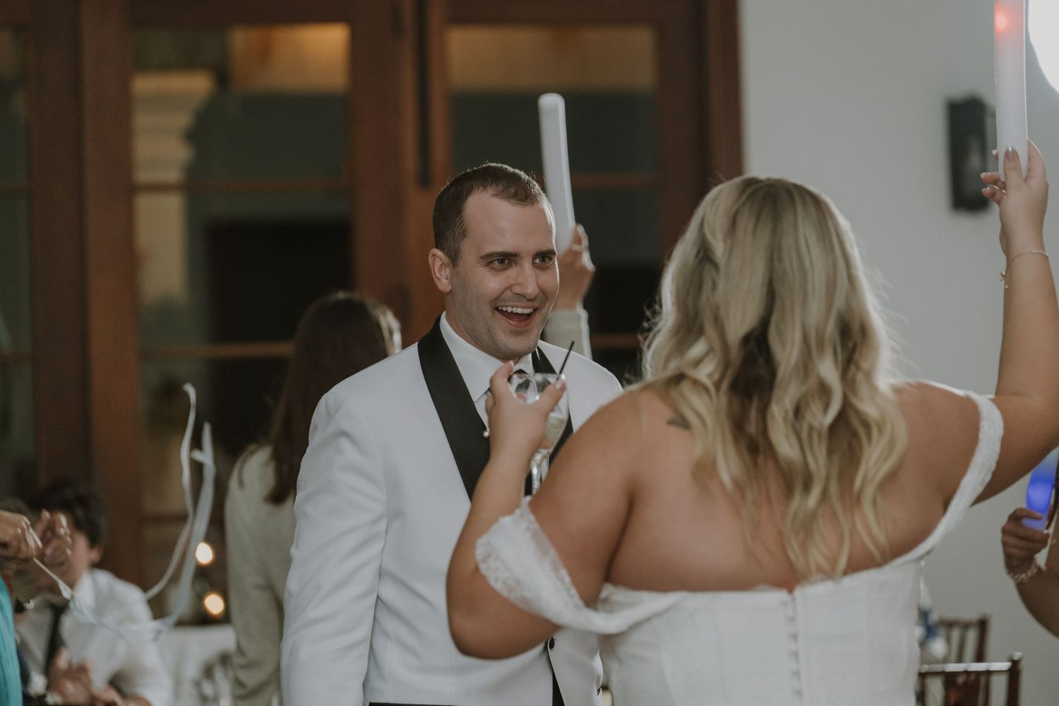 Groom in white jacket smiling at bride during a joyful wedding reception dance.
