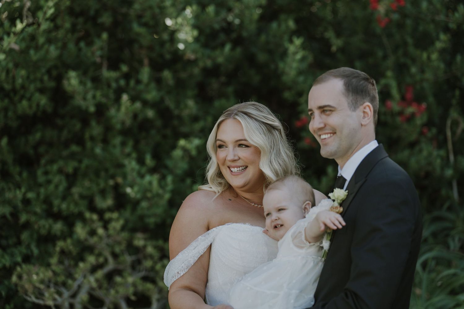 Bride and groom smiling outdoors holding a baby, surrounded by lush green foliage on their wedding day.