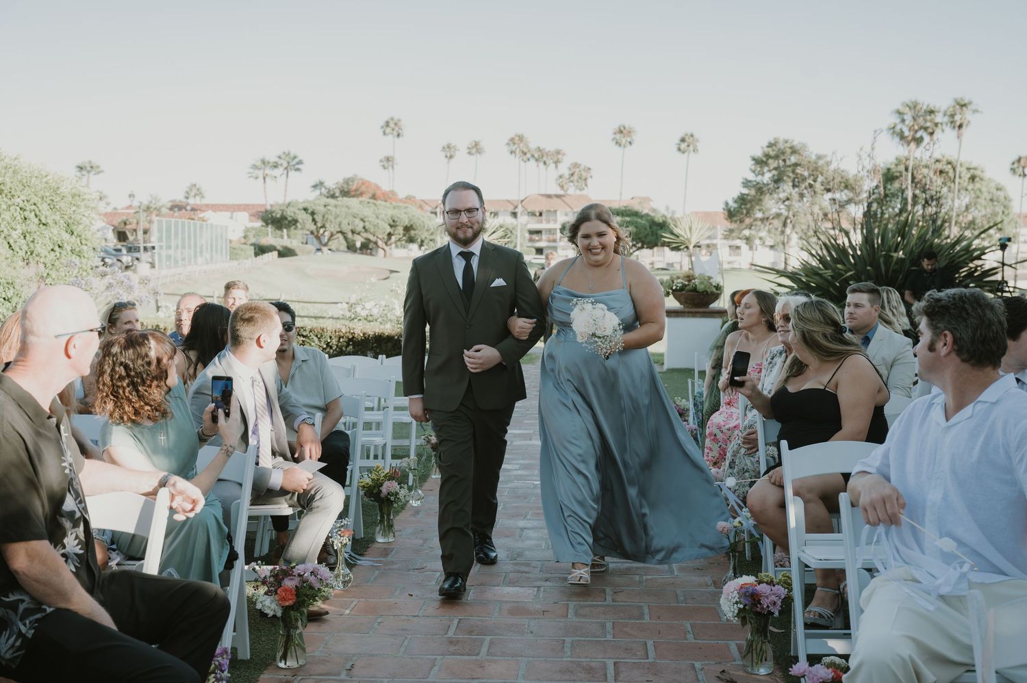 Bride and groom walk down the aisle at an outdoor wedding ceremony with guests seated on white chairs.