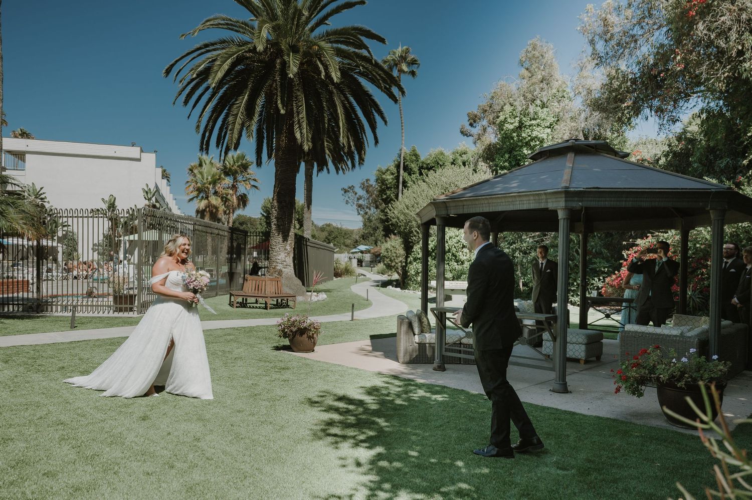 Bride and groom share a first look moment in a lush garden with a gazebo and palm trees on a sunny wedding day.