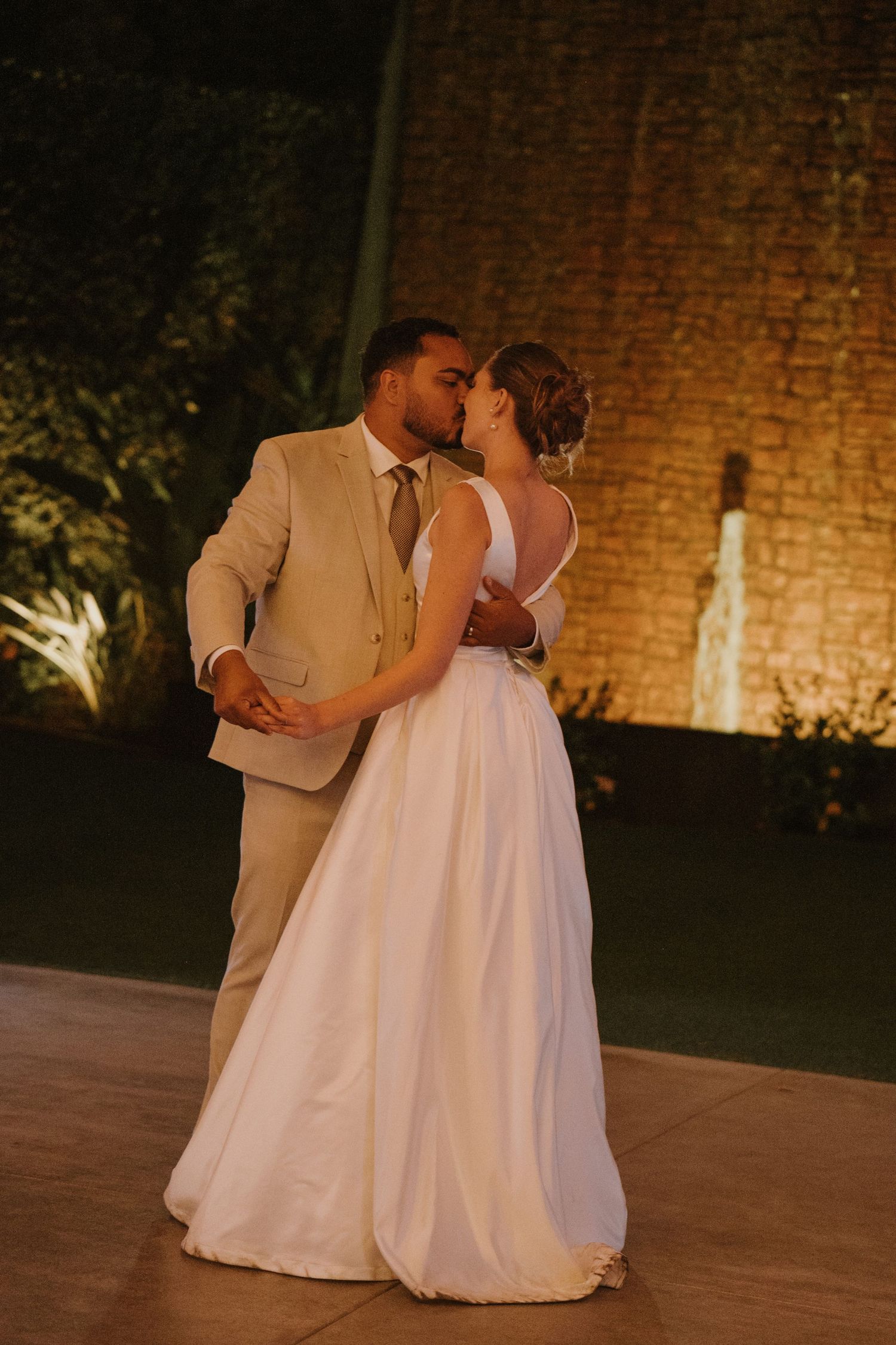 Newlyweds share a romantic kiss while dancing outdoors at night, bride in flowing white gown and groom in tan suit.