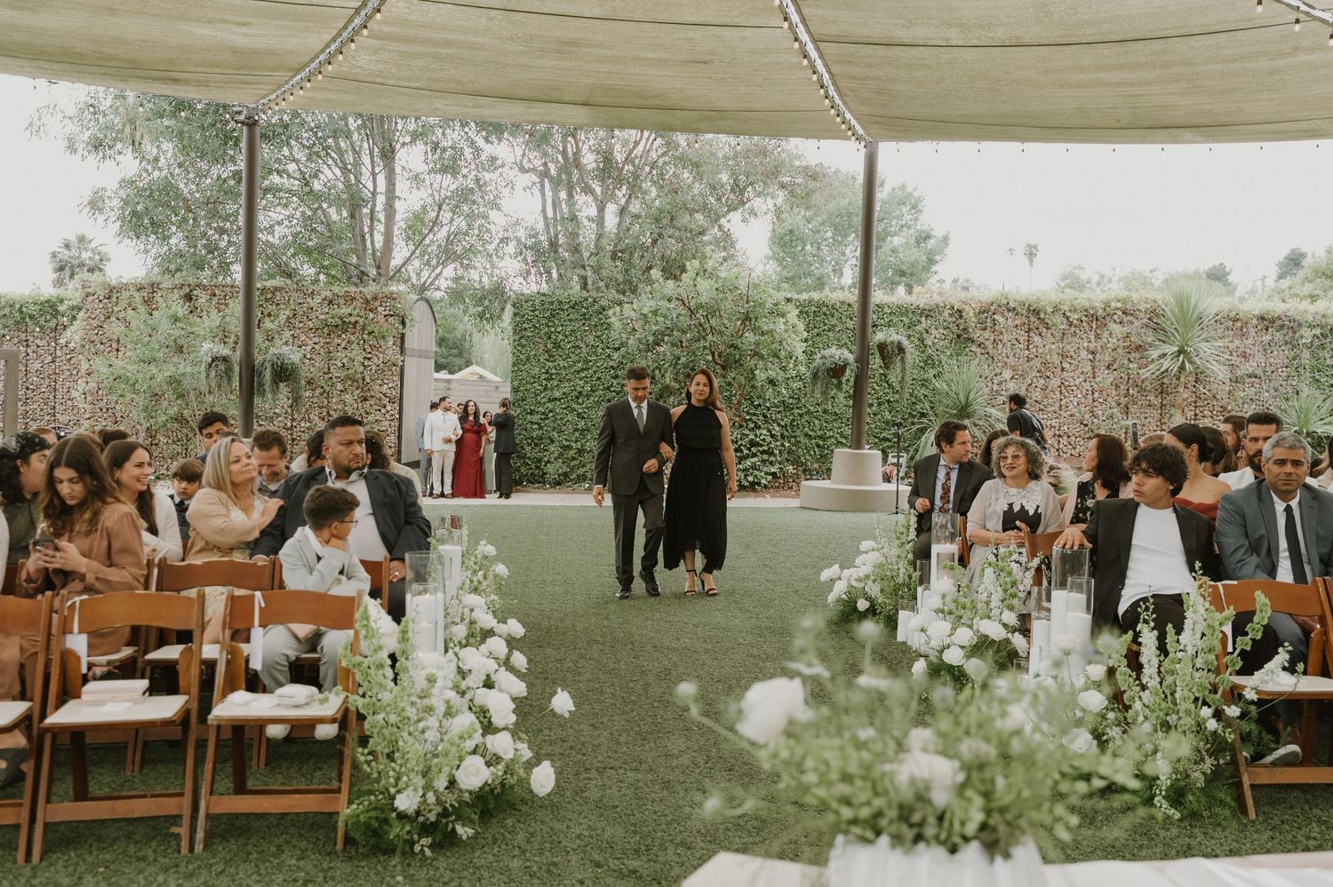 Outdoor wedding ceremony under a white tent with floral aisle decor and guests seated on wooden chairs.