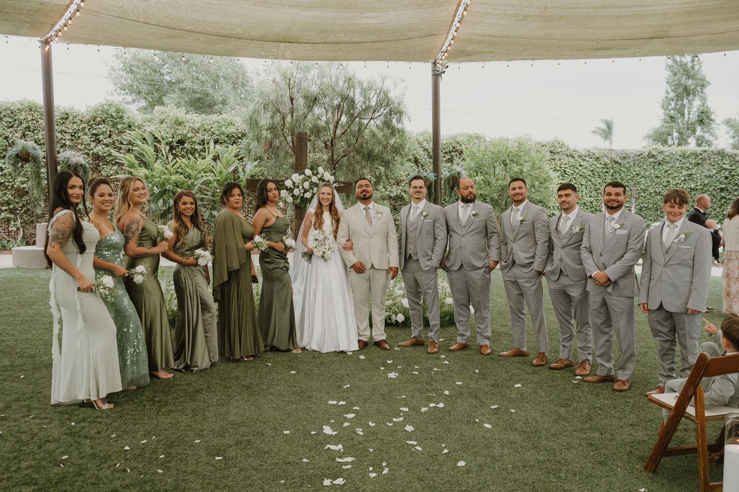 Wedding party poses together outdoors, bridesmaids in sage green dresses and groomsmen in gray suits.