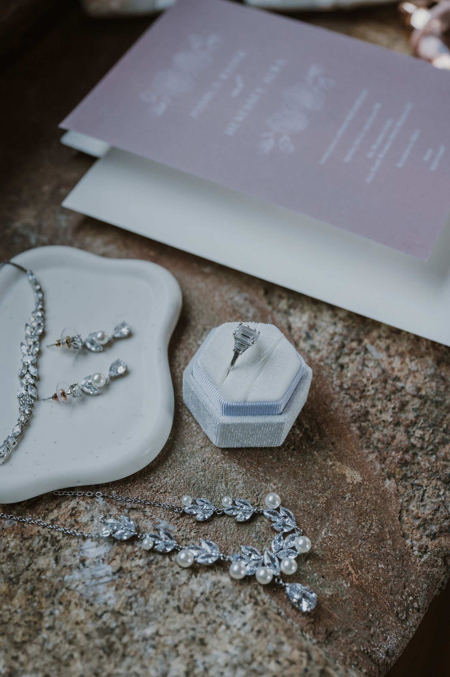 Elegant bridal jewelry set with crystal necklace, earrings, hair vine, and diamond ring in velvet box beside lavender wedding invitation.