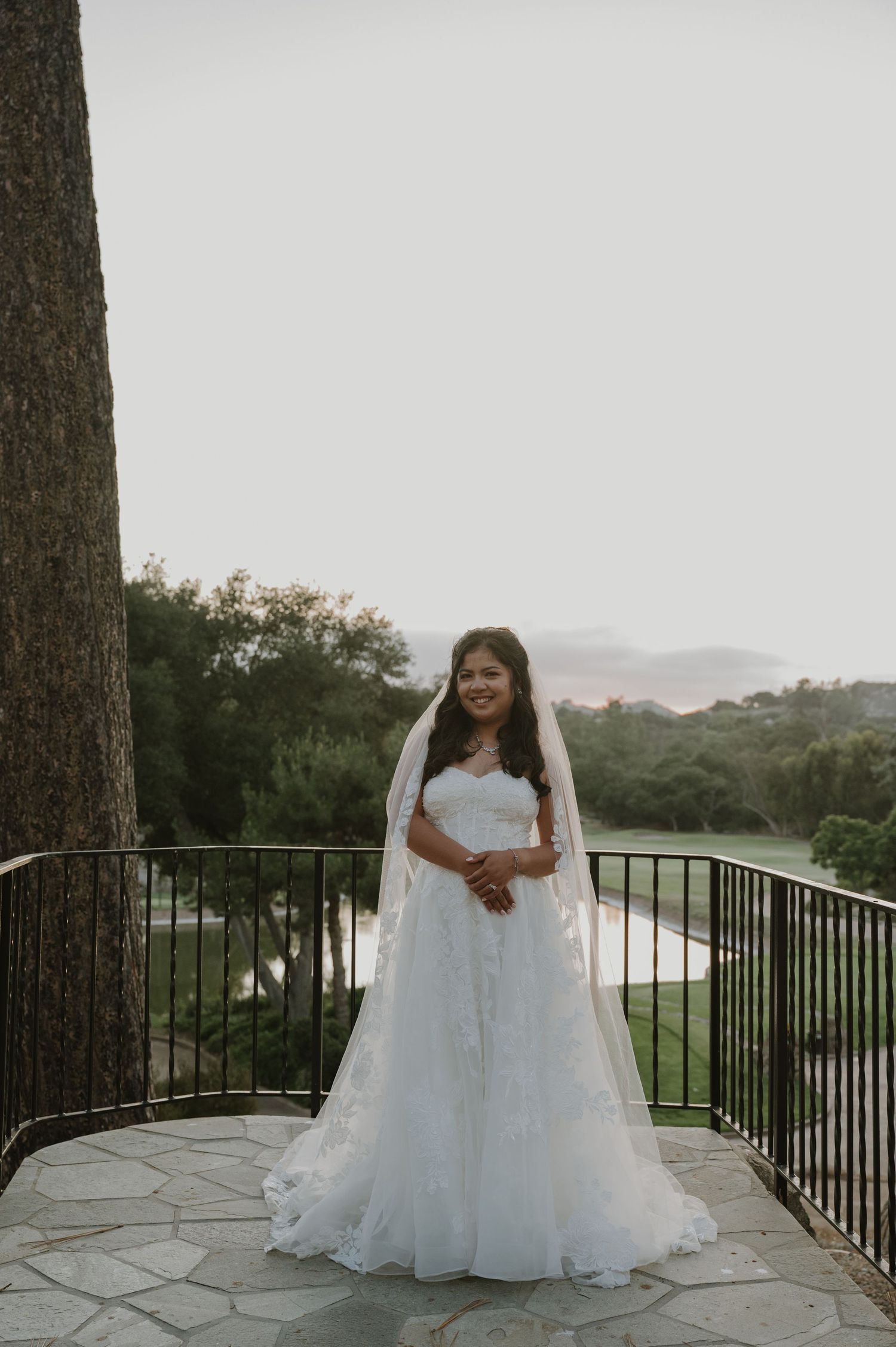 Bride in white gown and veil stands on stone balcony overlooking lush greenery and hills.