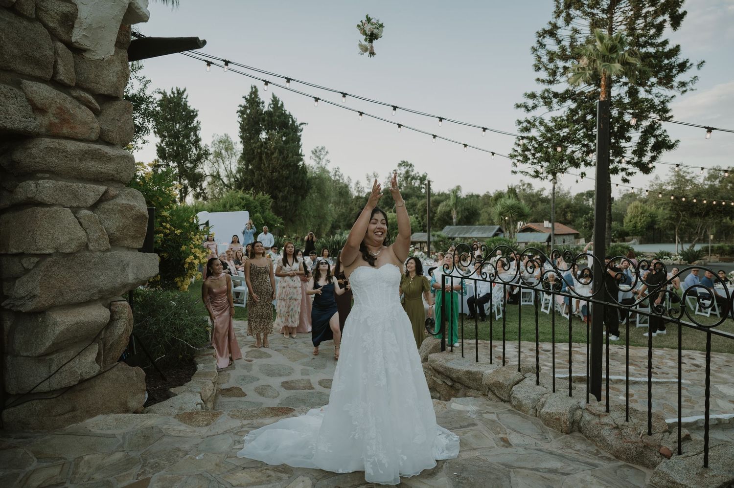 Bride in white strapless gown tossing bouquet to guests at outdoor wedding reception with string lights.
