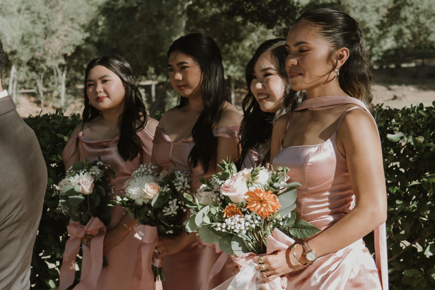 Four bridesmaids in blush pink dresses holding floral bouquets with roses and greenery at an outdoor wedding ceremony.