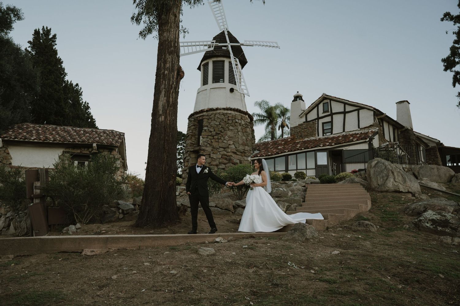 Wedding couple stands near historic windmill and rustic building during golden hour.
