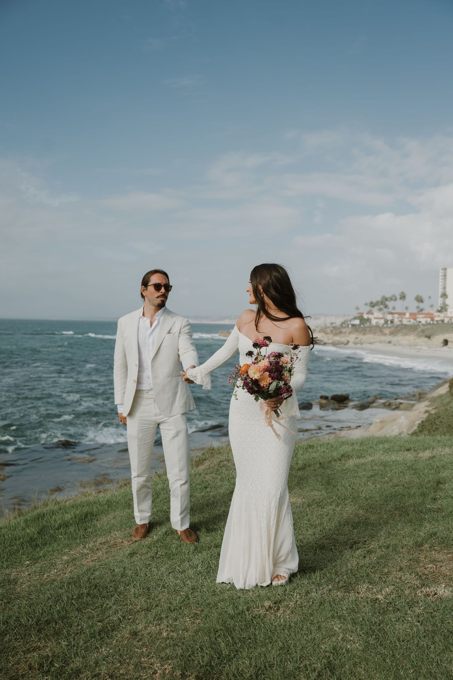 A wedding couple walks and dances together on a grassy oceanside cliff.