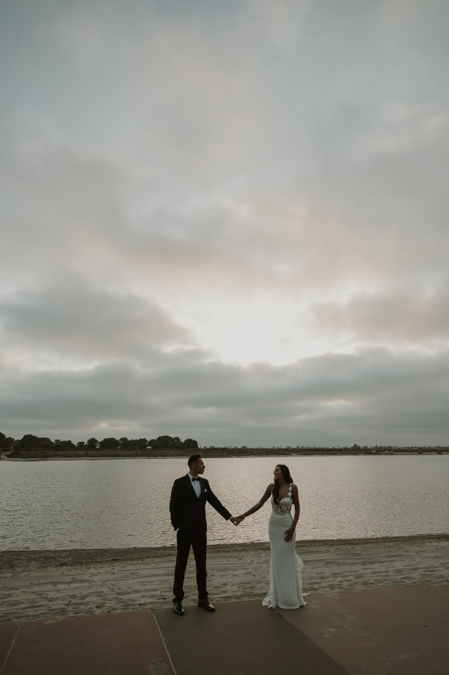 A couple in wedding attire holds hands on a beach during an overcast day with water and distant shoreline visible.