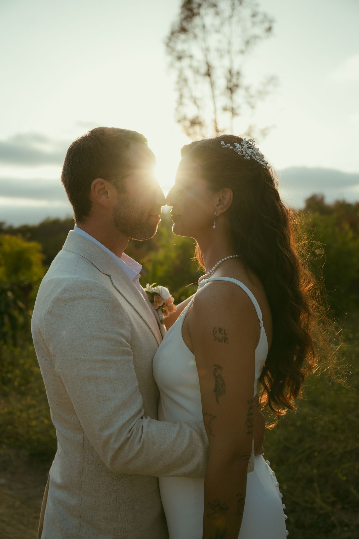 Bride and groom share a romantic golden hour moment outdoors, sun glowing behind them, her tattoo and tiara visible.