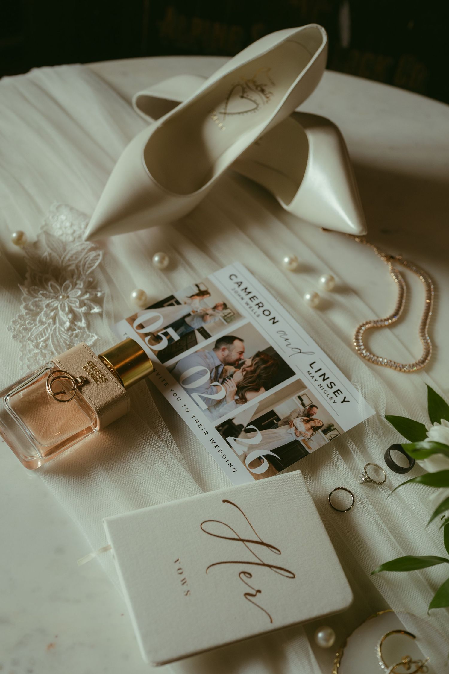 Elegant bridal flat lay featuring white satin heels, pearl jewelry, perfume bottle, wedding invitation, and a gold-lettered gift box.
