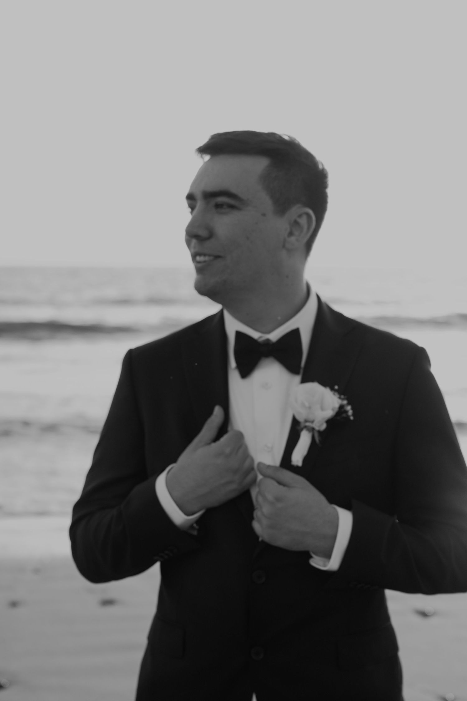 Groom in black tuxedo with bow tie and boutonniere smiling on beach in black and white wedding photo.