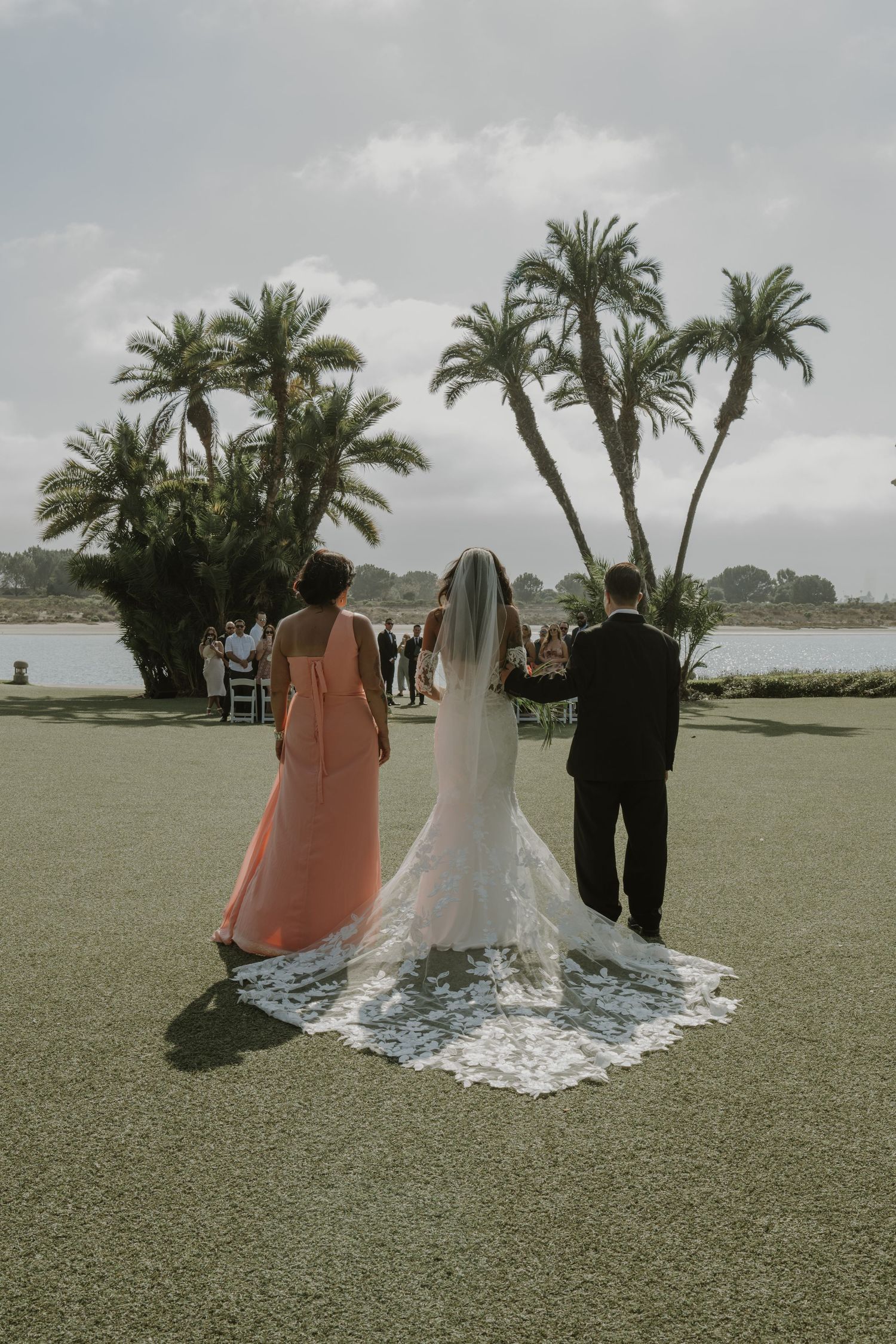 Wedding ceremony attendees stand on a grassy lawn with palm trees and waterfront views.