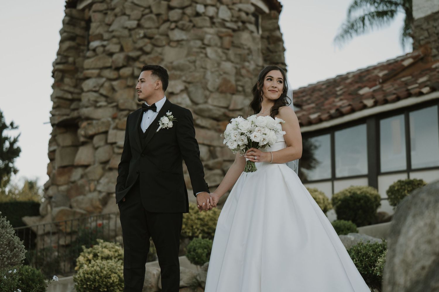 Wedding couple posing outside a rustic stone building with elegant landscaping.