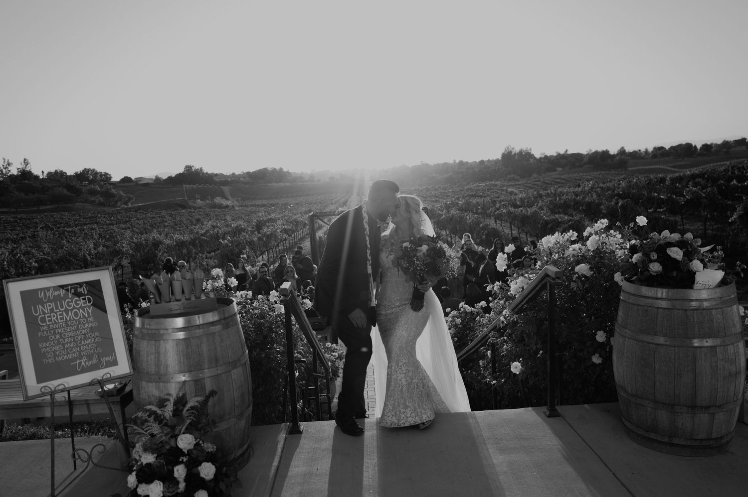 Black and white photo of newlyweds walking down decorated vineyard path at sunset.