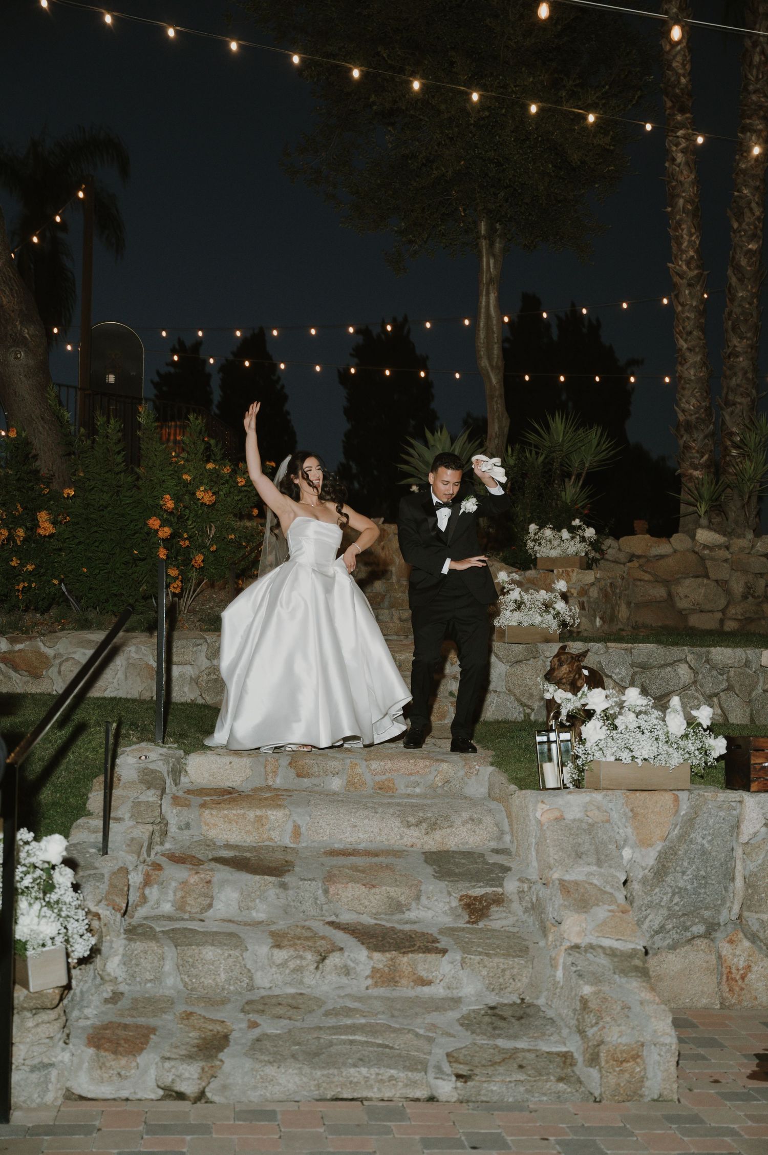 Evening outdoor wedding celebration with string lights and stone wall decorations.