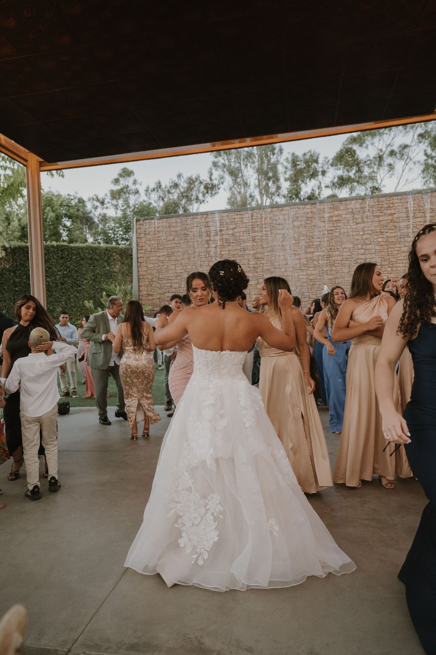 Bride in a white floral gown dancing with guests at an outdoor wedding reception.