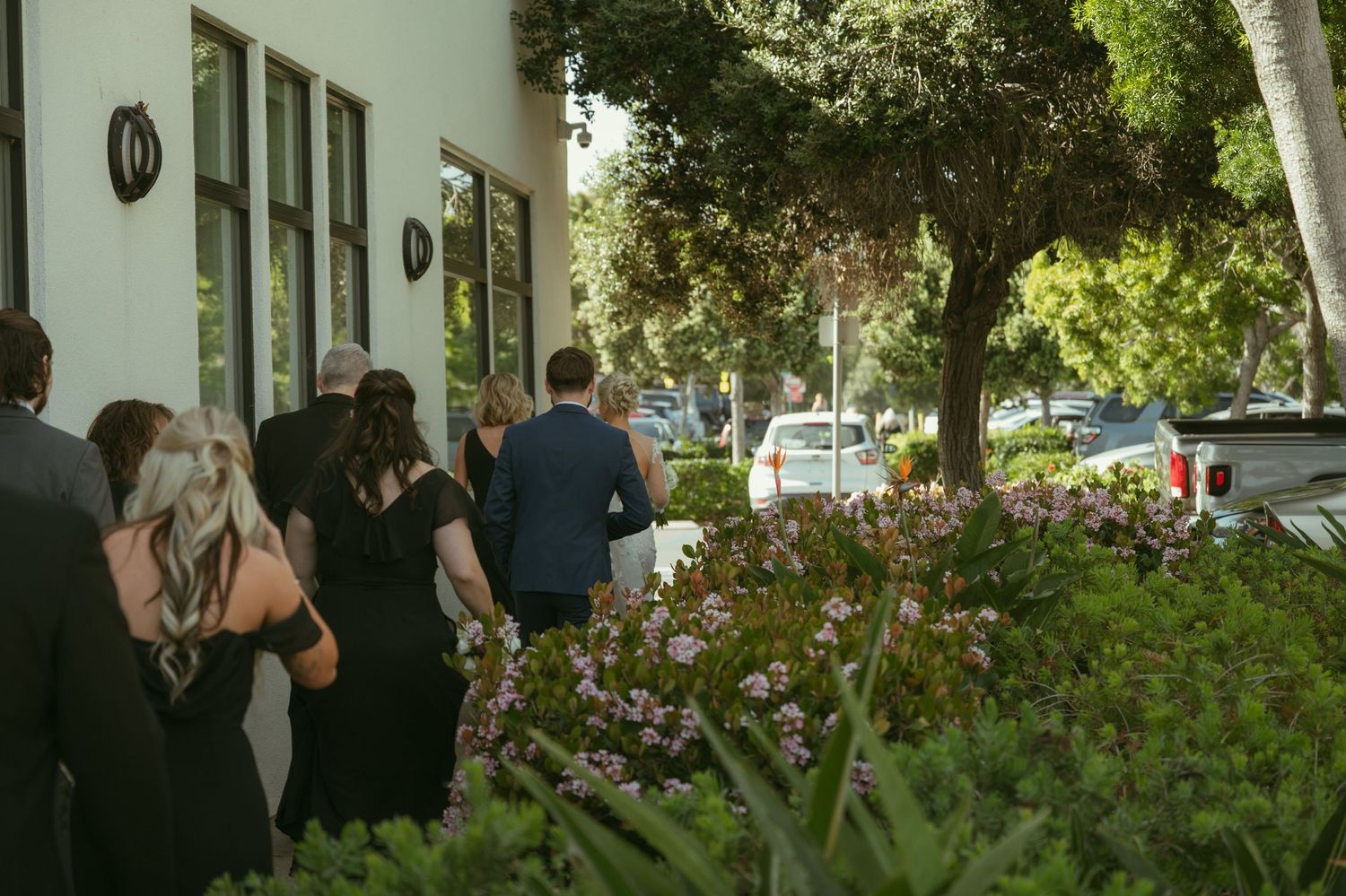 Guests in formal attire walk along a flower-lined path toward a building entrance surrounded by lush green trees.
