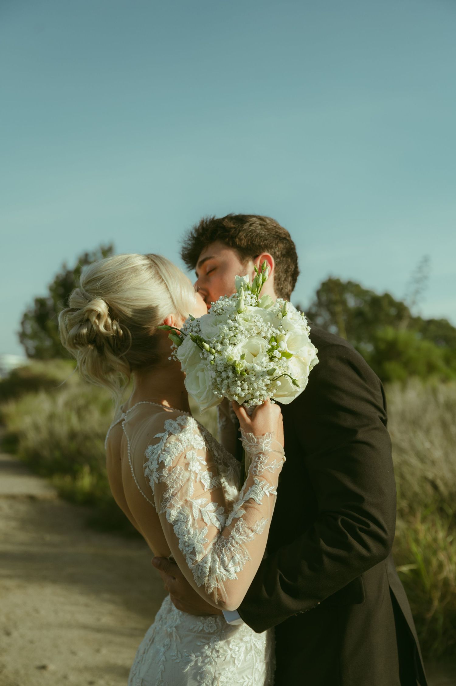 Bride and groom share a kiss behind a white floral bouquet outdoors.