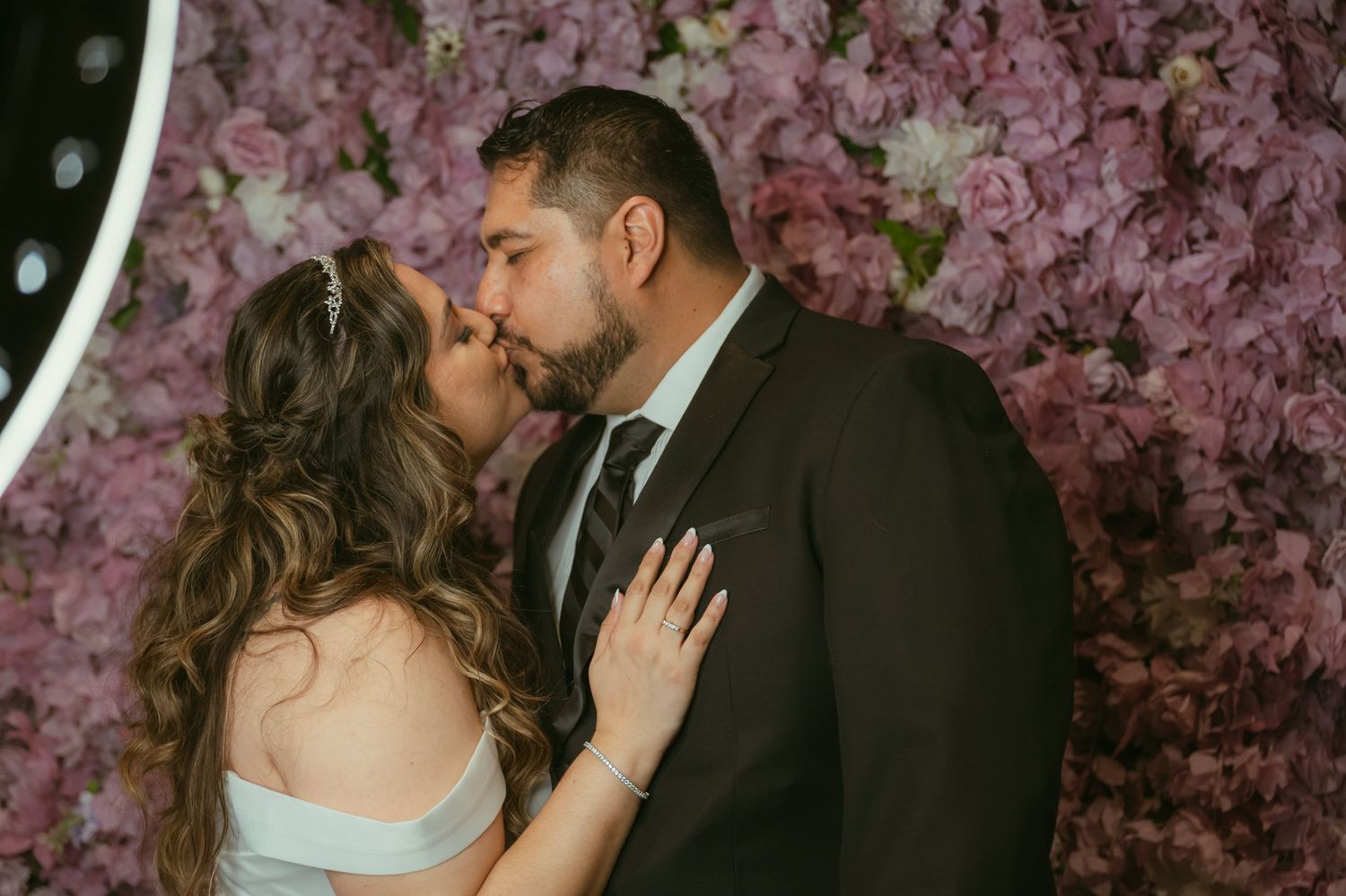 Couple sharing a romantic kiss in front of a lush pink floral wall at their wedding reception.