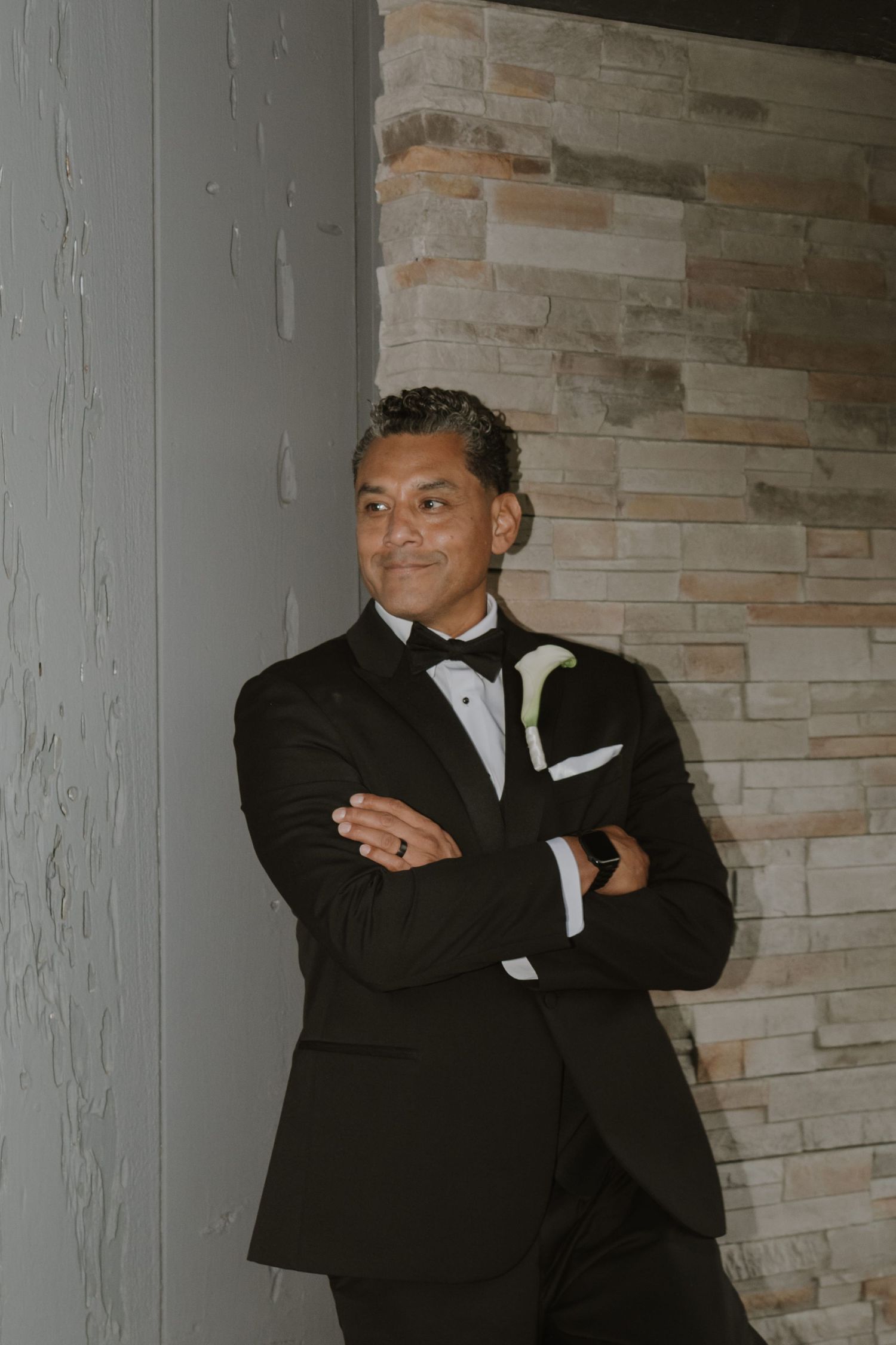 A man in a black tuxedo with bow tie and boutonniere stands arms crossed against a stone wall.