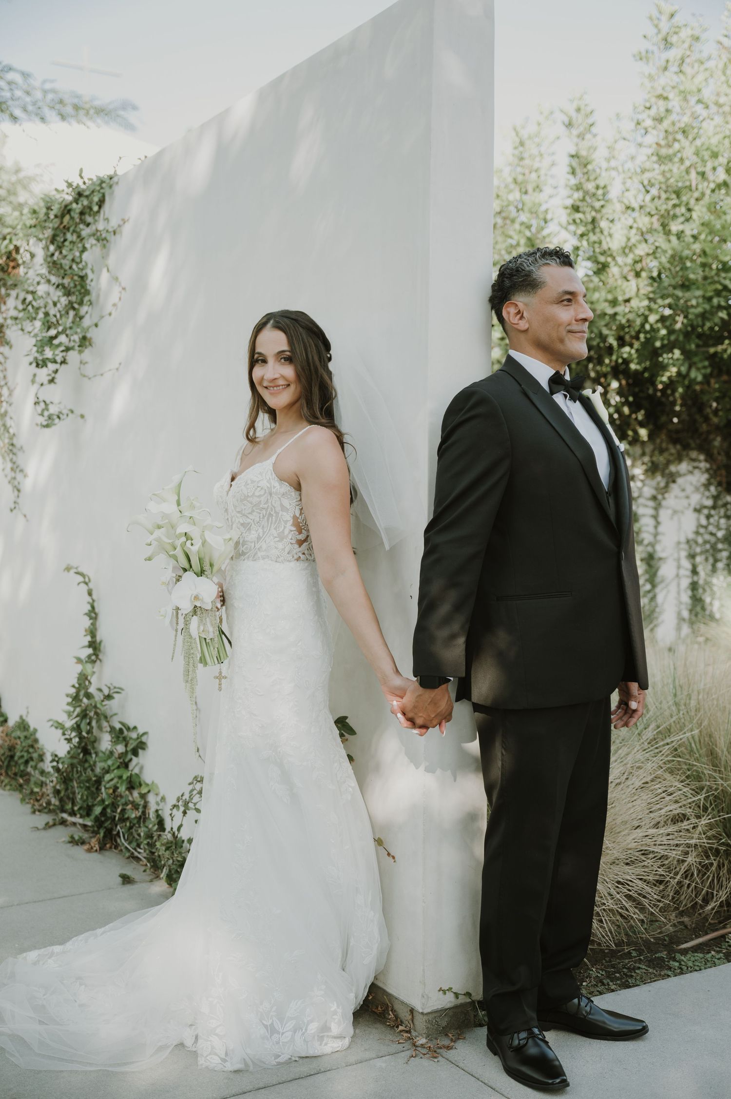 Bride in lace gown holds bouquet while groom in black tuxedo holds her hand against a white wall.