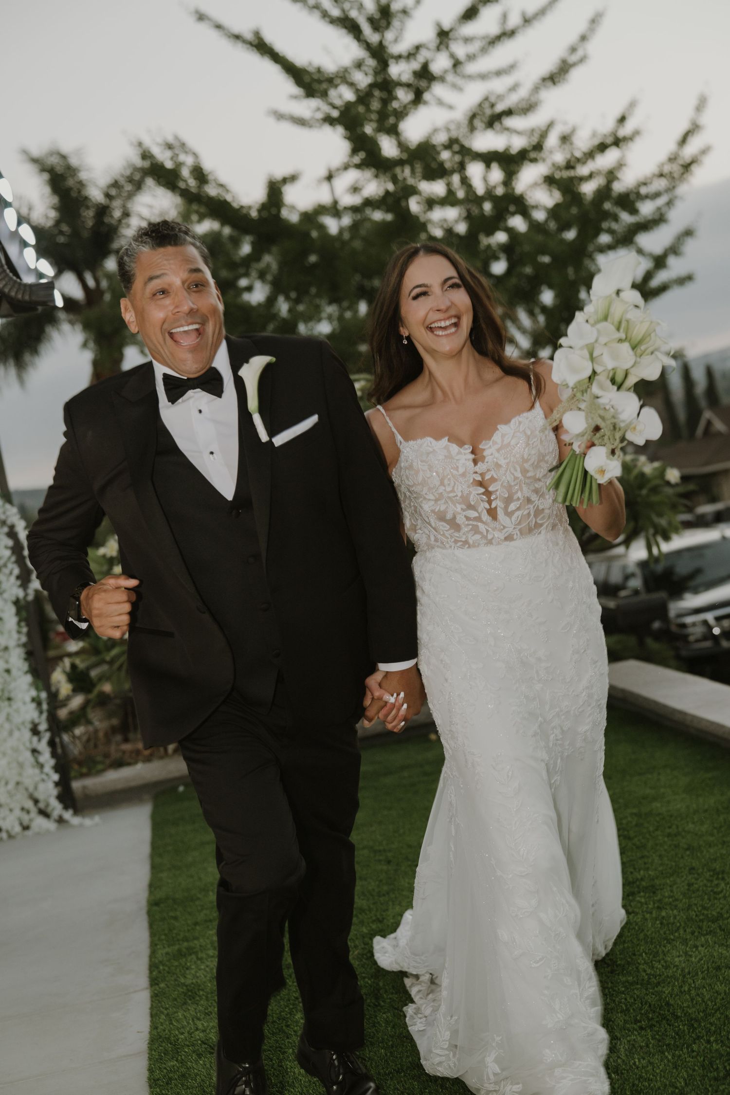 Joyful newlyweds walk hand in hand, bride holding white bouquet in elegant lace wedding gown.