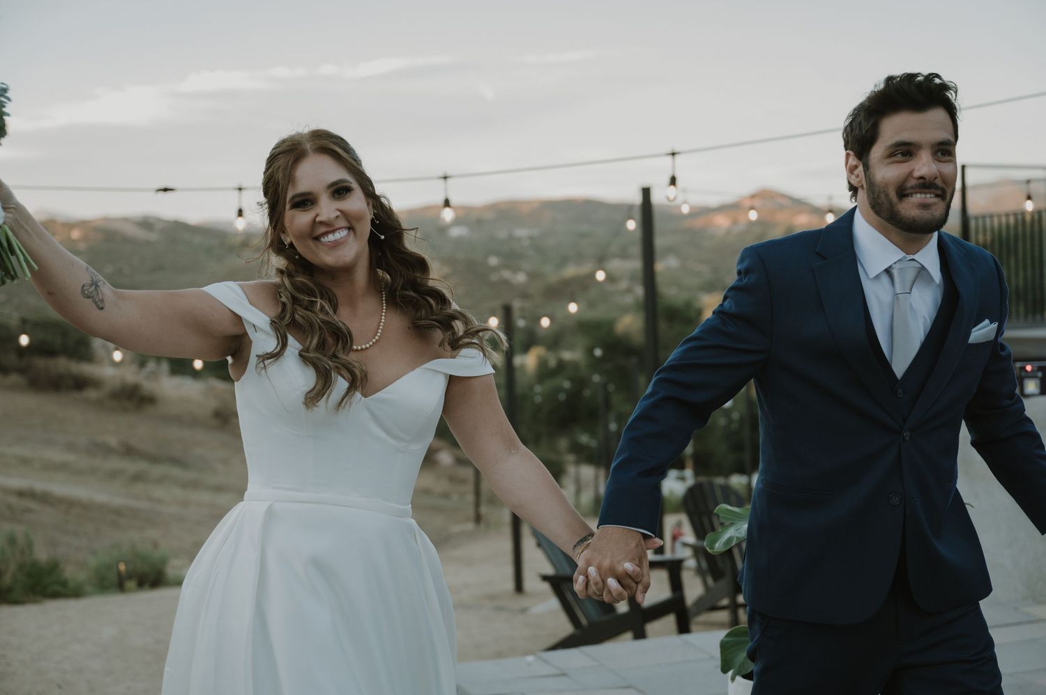 Happy newlyweds holding hands outdoors at a rustic wedding venue at dusk, bride in white gown and groom in navy suit.