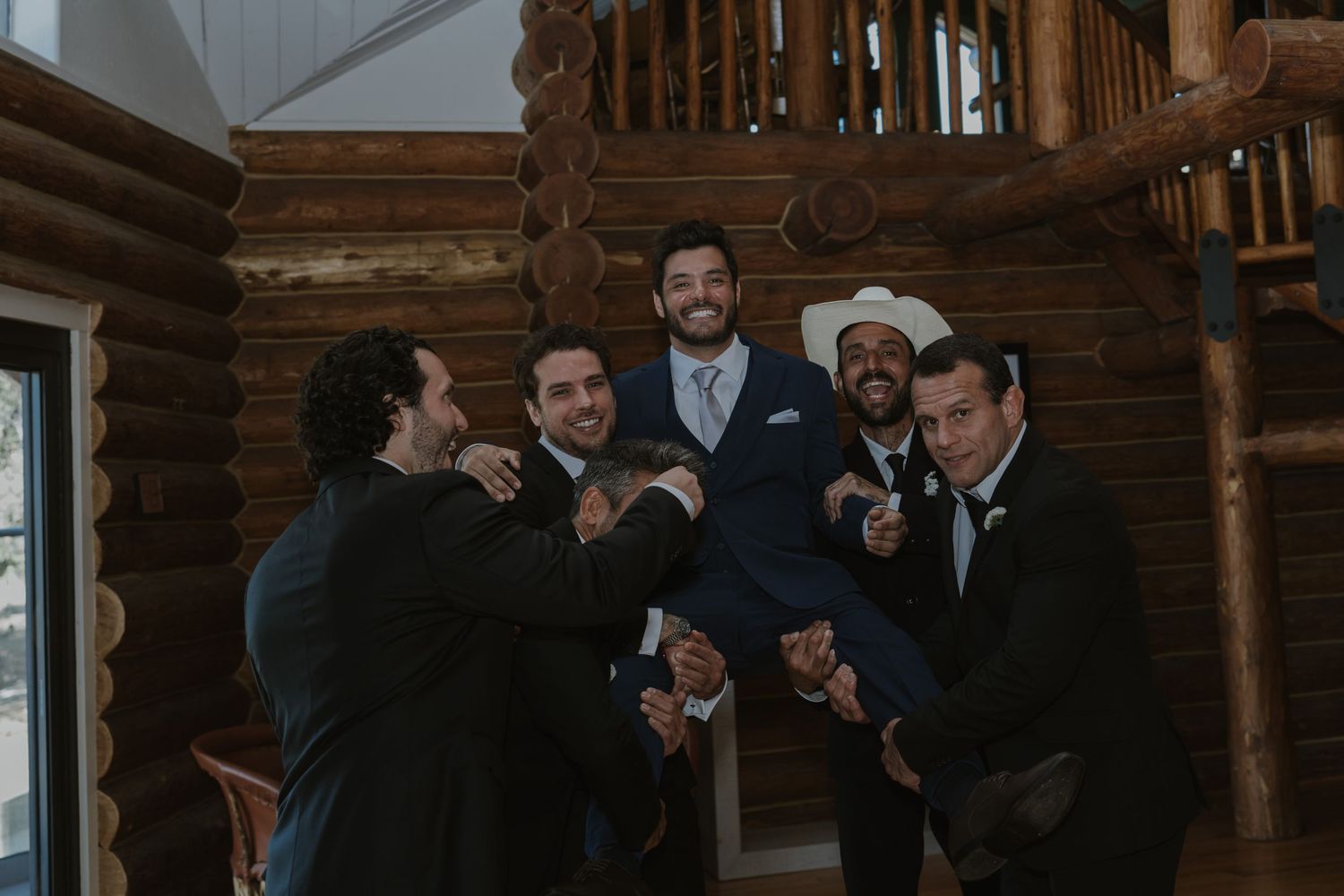 Groomsmen laughing and lifting the groom in a log cabin wedding venue before the ceremony.