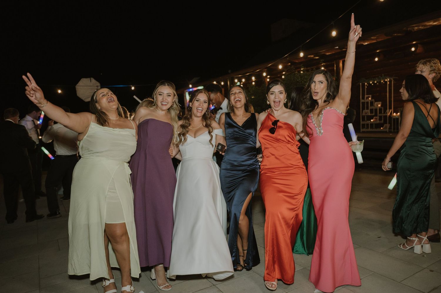 Bride celebrating with six bridesmaids in colorful formal gowns under string lights at outdoor wedding.