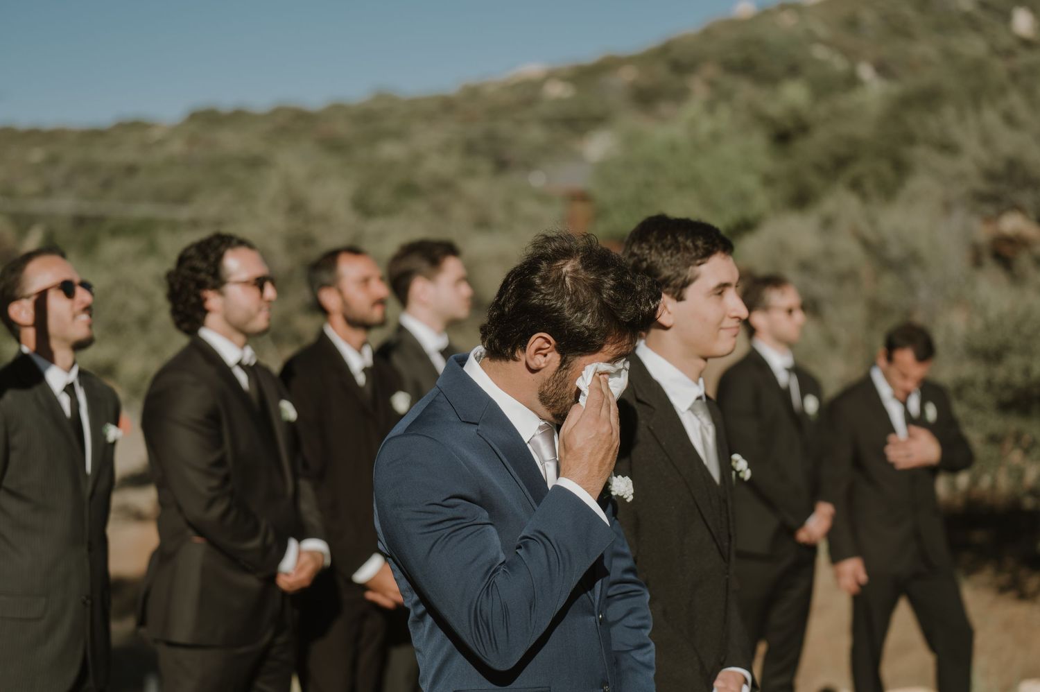 Emotional groom wiping tears with groomsmen standing in line outdoors at wedding celebration.