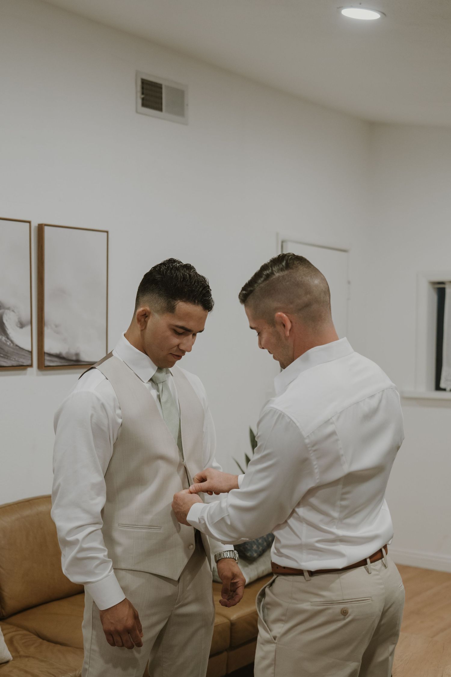 Groomsman helping groom adjust boutonniere on tan vest during wedding preparations in a modern white room.