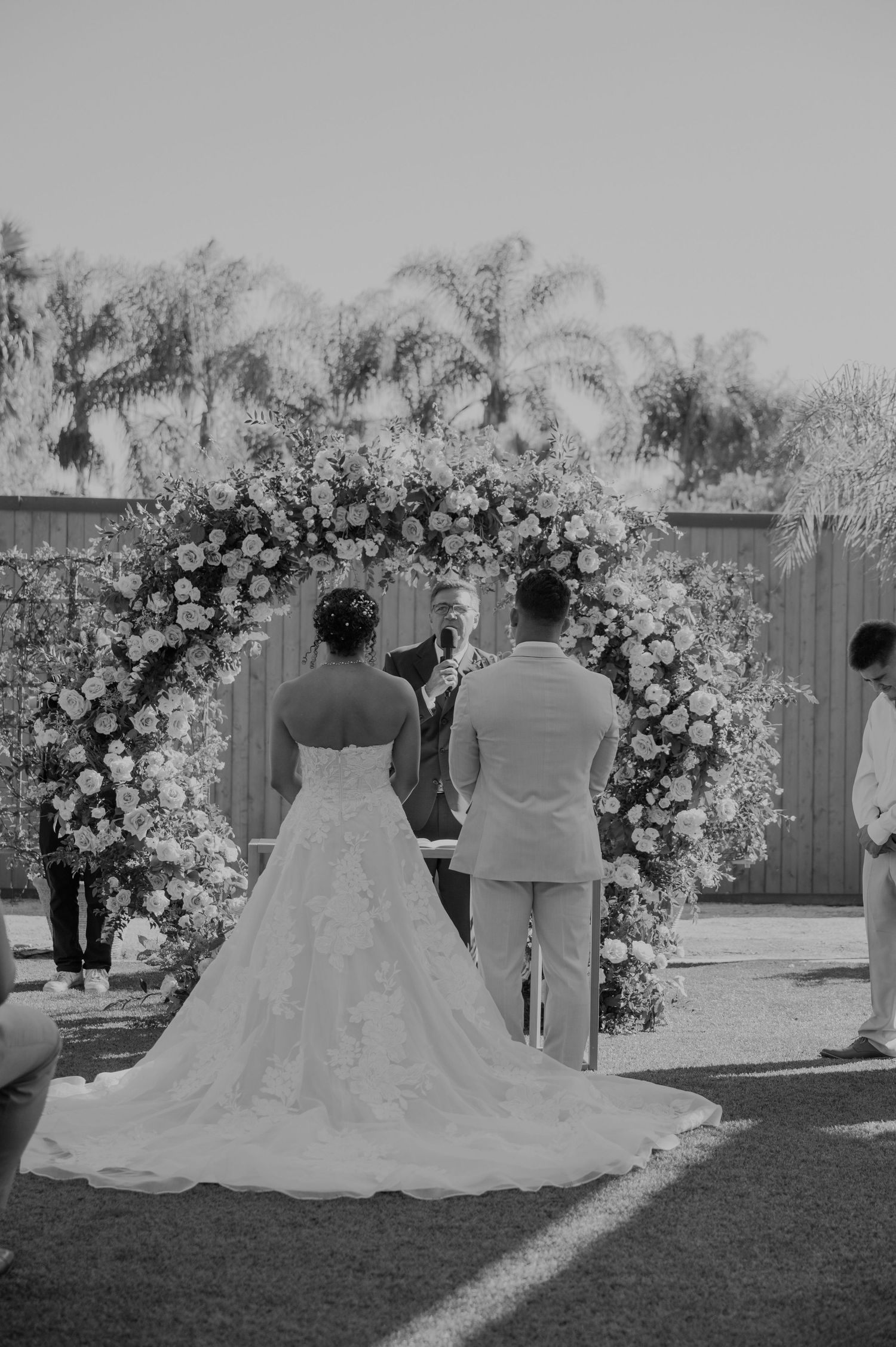 Black and white outdoor wedding ceremony with couple at floral arch, officiant, and palm trees in background.