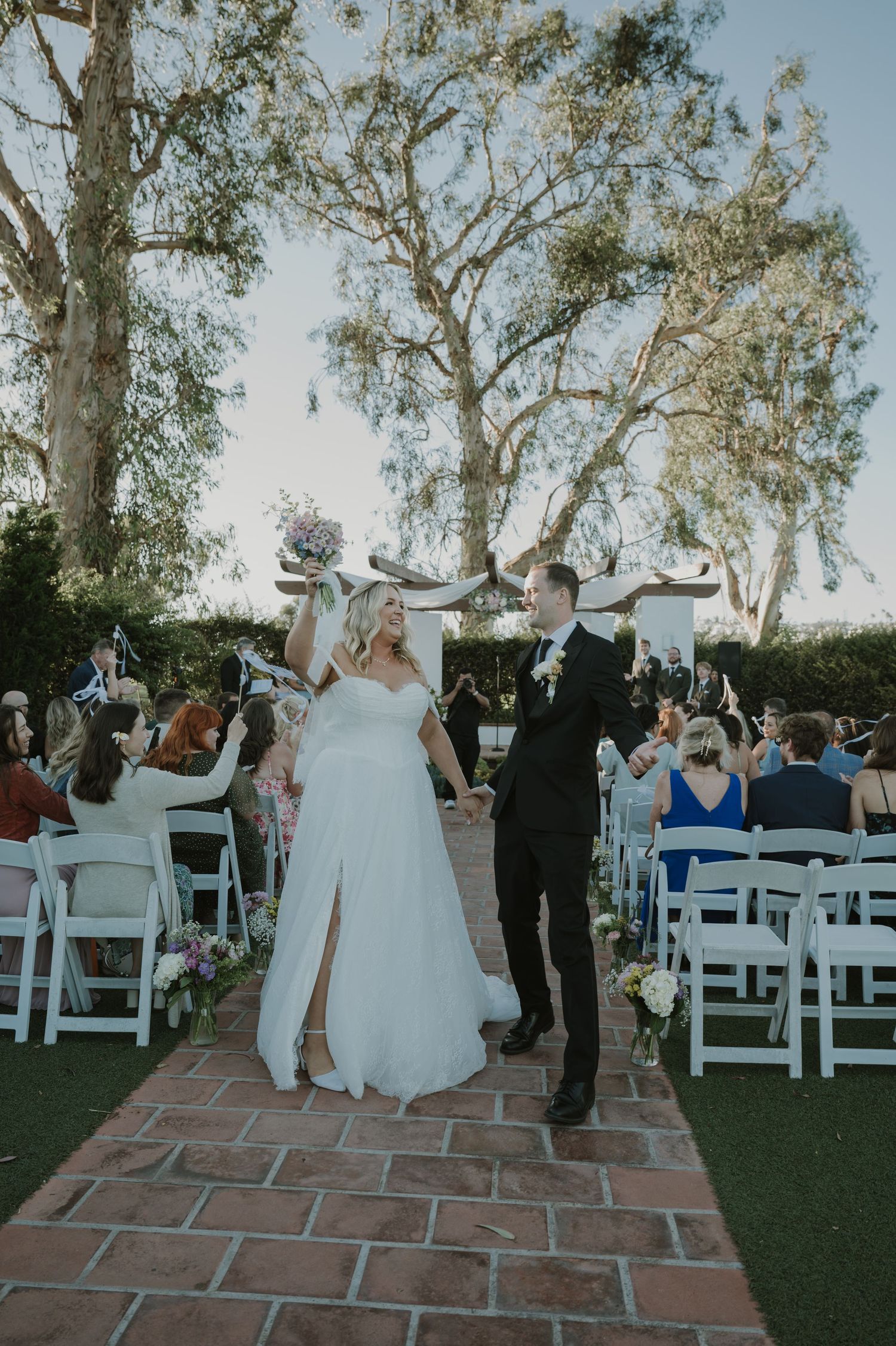 Joyful newlyweds raise hands celebrating as they walk back down aisle after outdoor garden wedding ceremony.