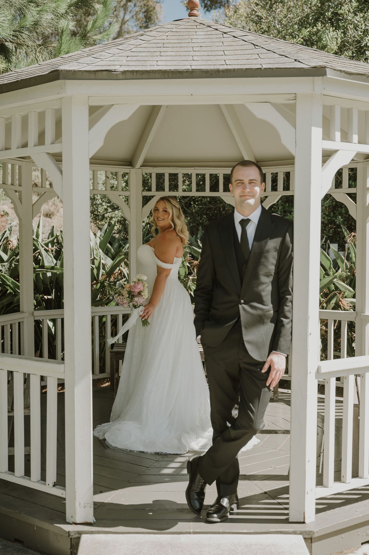 Groom in black suit leans on white gazebo post as bride in white gown stands behind him.