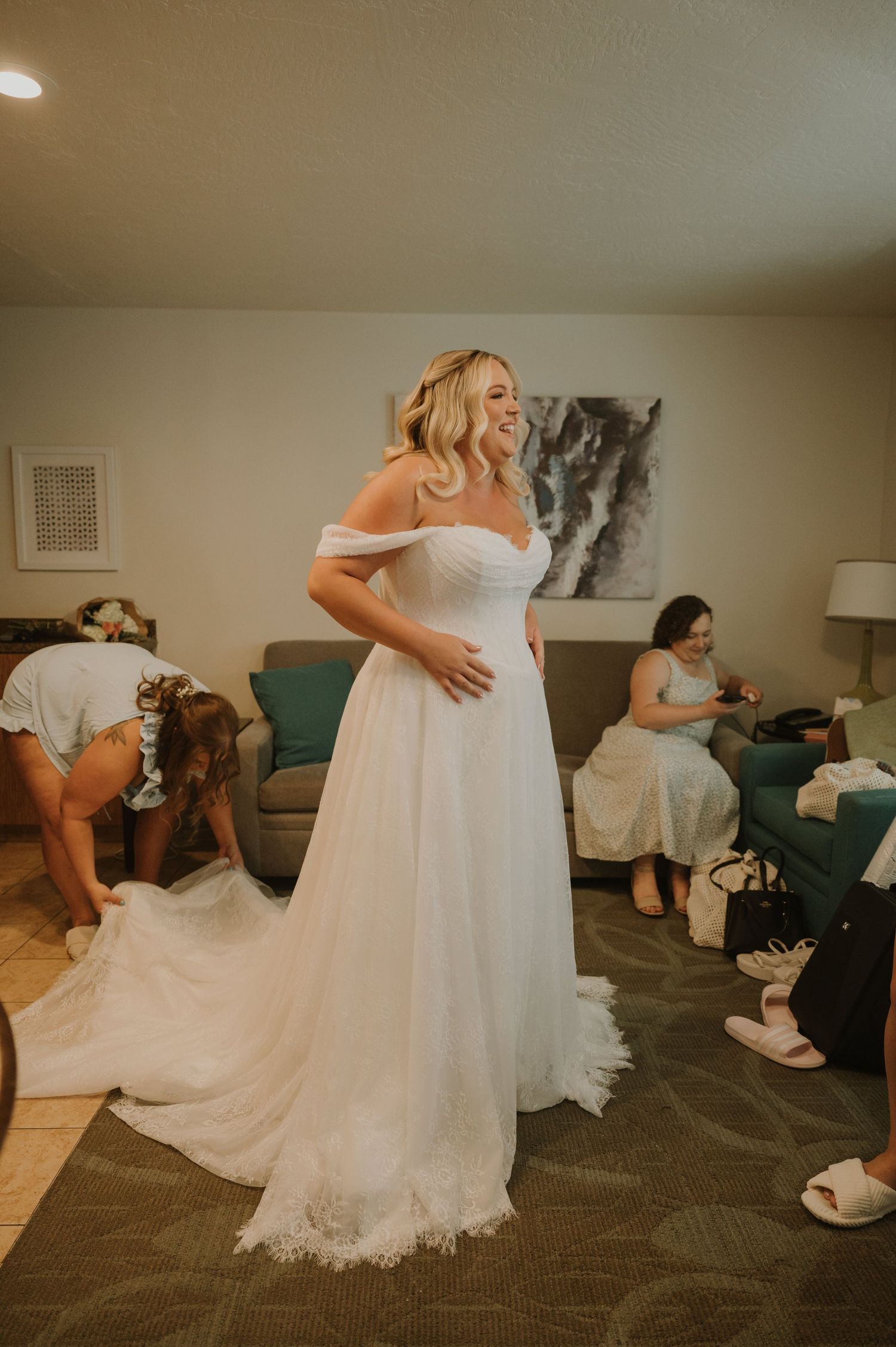 Bride in elegant off-shoulder white wedding gown with floral hem train getting ready with bridesmaids in hotel room.