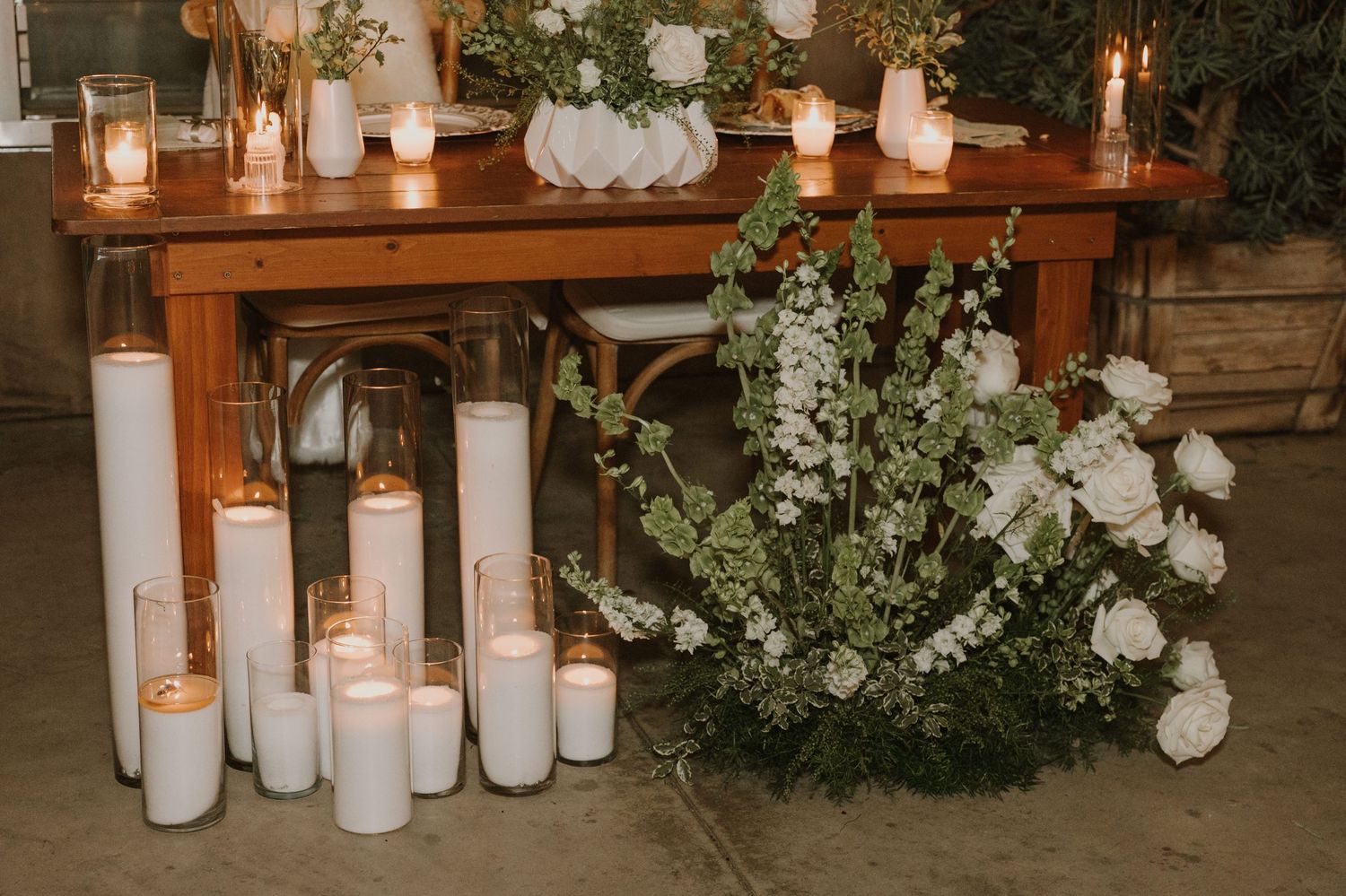 Rustic wooden wedding table decorated with white pillar candles and lush white floral arrangements on the floor.