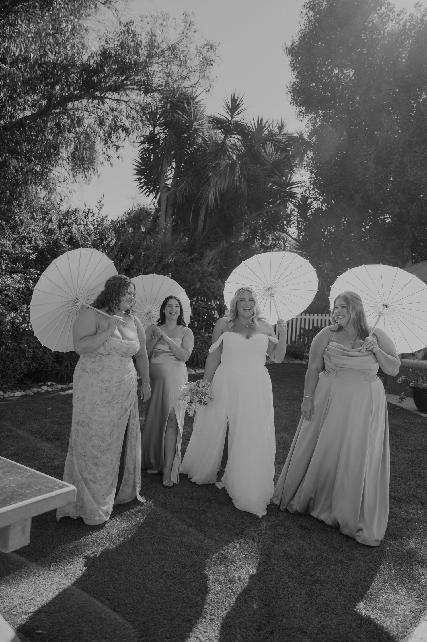 Black and white photo of bride and three bridesmaids holding white parasols walking outdoors at wedding.