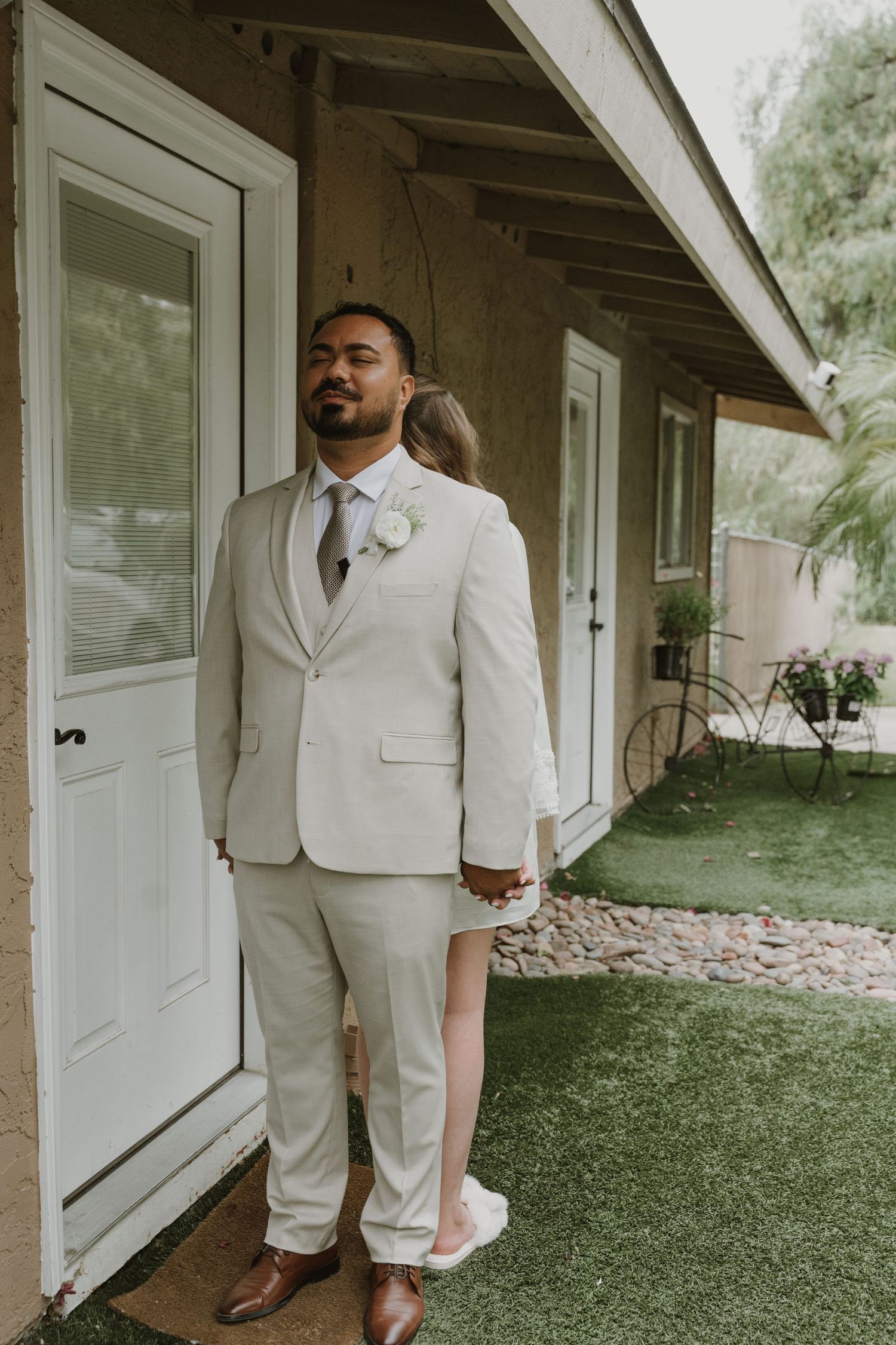 Groom in beige suit with white boutonniere stands confidently outside while holding bride's hand behind him.
