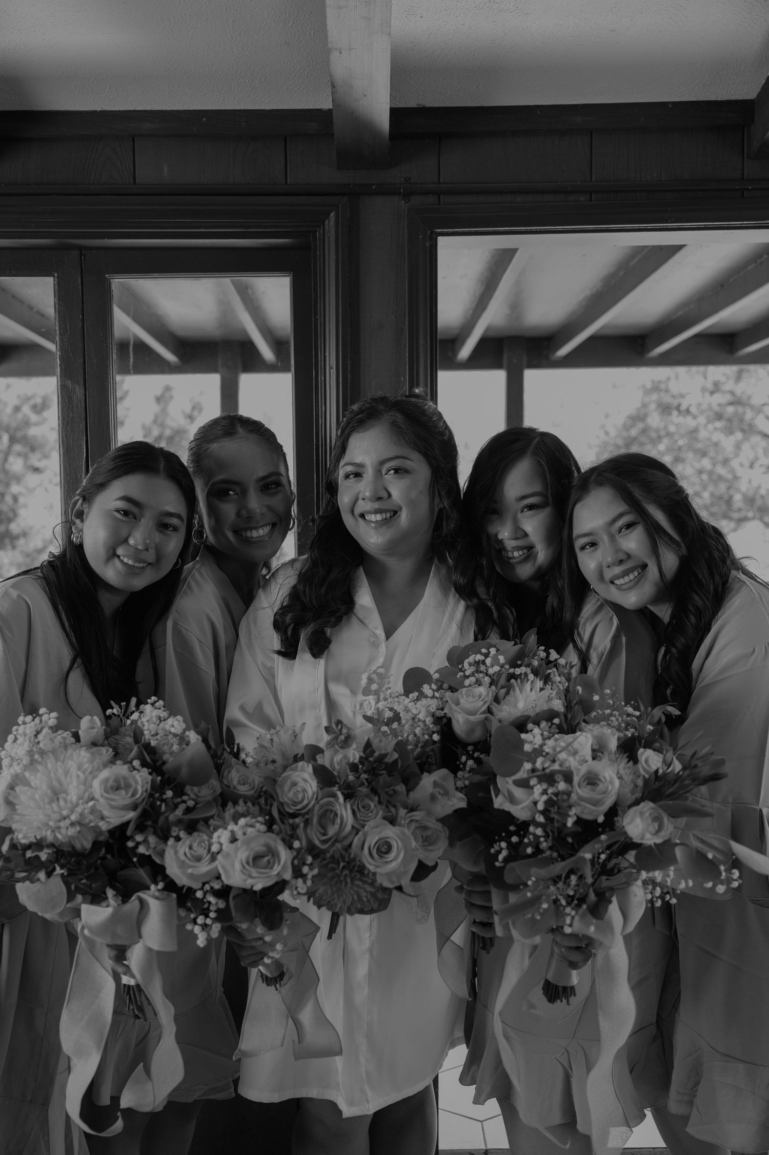 Bride and four bridesmaids in robes holding floral bouquets, smiling in black and white photo.