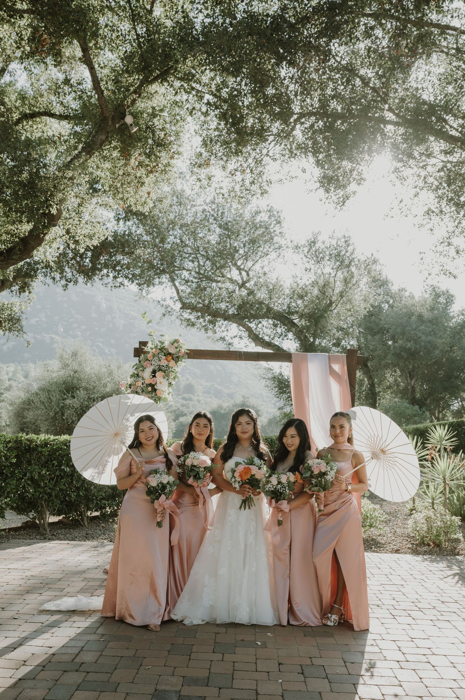 Bride in white gown with four bridesmaids in blush pink dresses holding bouquets and parasols outdoors.